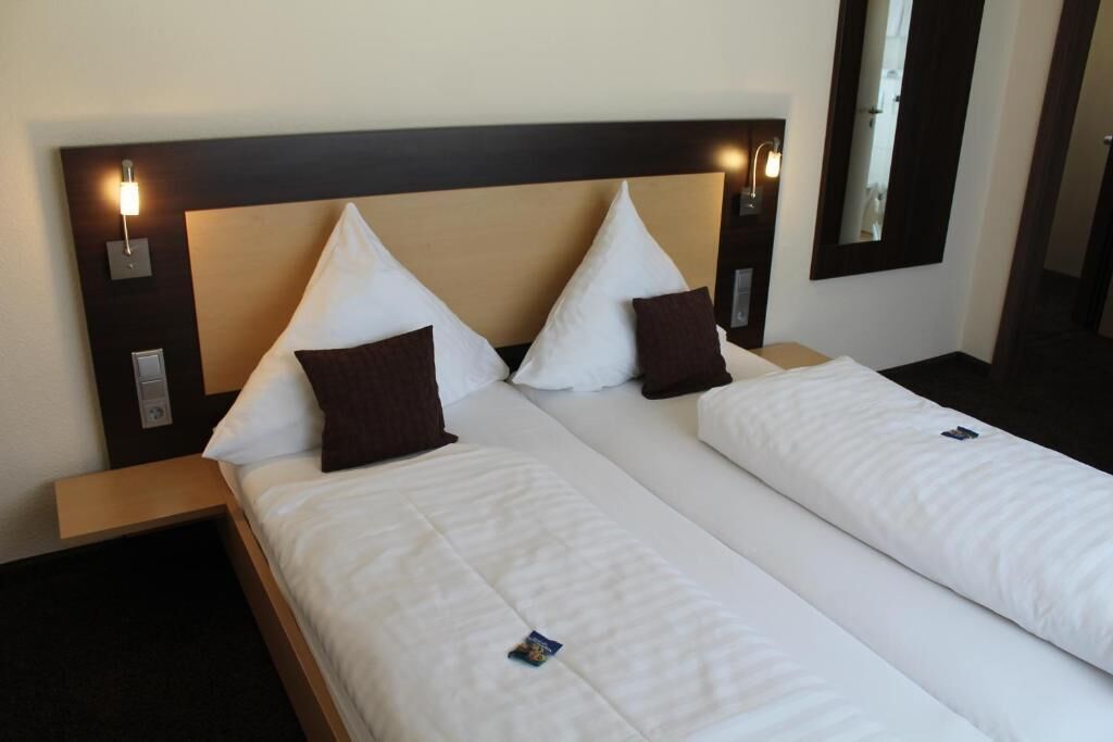 A hotel room with two beds, each with white bedding and brown pillows, a wooden headboard, bedside lamps, and a mirror on the wall.