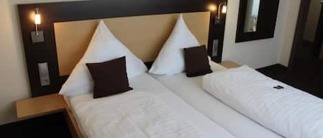A hotel room with two beds, each with white bedding and brown pillows, a wooden headboard, bedside lamps, and a mirror on the wall.
