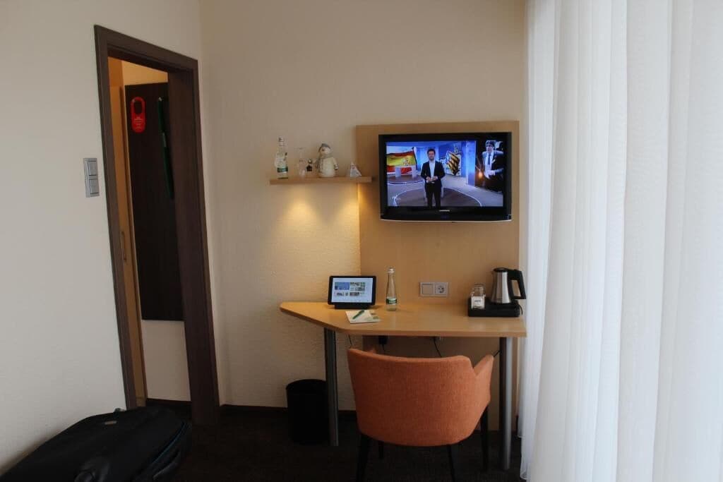 Ein Hotelzimmer mit Schreibtisch, Wandfernseher, Stuhl und Koffer.