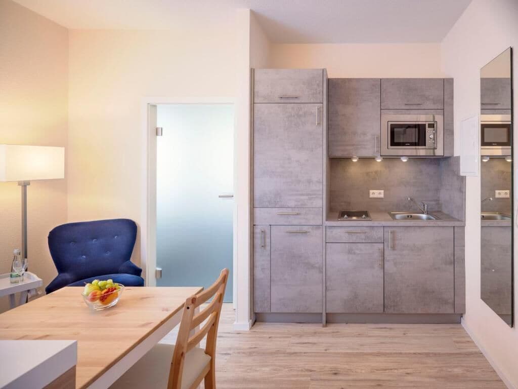 Een moderne keuken met grijze kastjes, een magnetron en een spoelbak. Er is een eethoek met een tafel en stoelen, en een blauwe stoel.