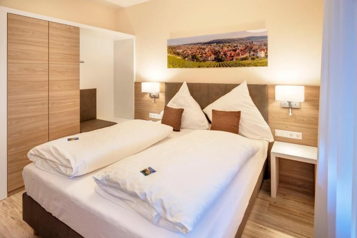 Una cama doble con sábanas blancas, dos mesitas de noche con lámparas, un armario de madera y un cuadro colgado en la pared que muestra un paisaje.