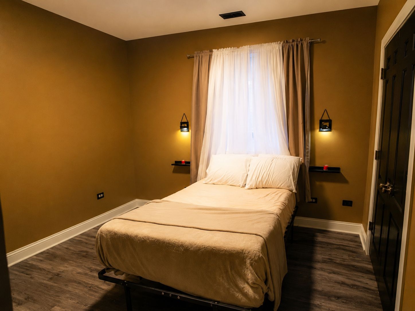 A single bed in a room with a window covered by sheer curtains, two wall-mounted lights, and a door on the right.