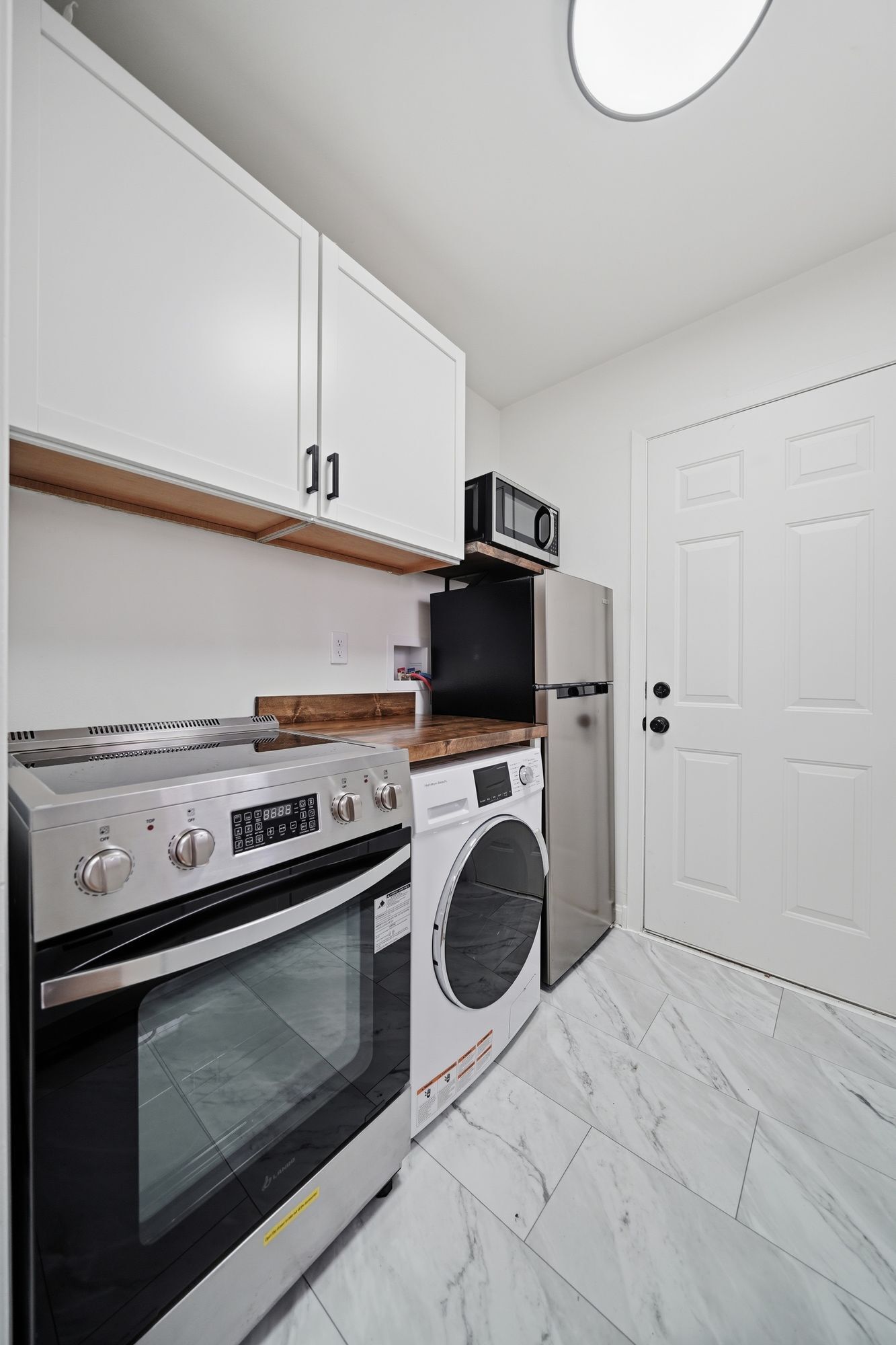 A kitchen with a built-in oven, microwave, and washing machine, a wooden countertop, and a white door.