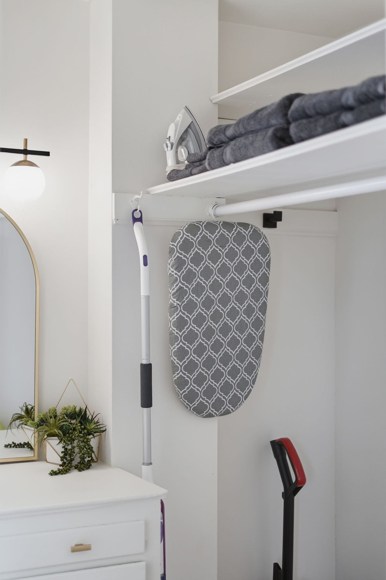 A bathroom with a white vanity, a hanging towel rack, a wall-mounted mirror, a floor lamp, a potted plant, and a red and black vacuum cleaner.