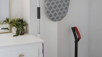 A bathroom with a white vanity, a hanging towel rack, a wall-mounted mirror, a floor lamp, a potted plant, and a red and black vacuum cleaner.