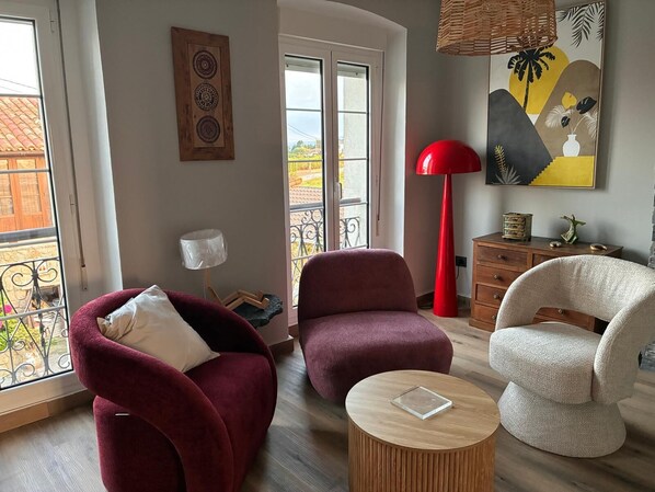 Una sala de estar con dos sillones, una mesita redonda de madera, una lámpara de pie roja, una lámpara blanca y un cuadro en la pared.