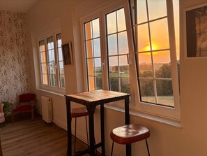 Ein Zimmer mit einem Holztisch, zwei Hocker, großen Fenstern mit Sonnenuntergangsblick und einer Heizung an der Wand.