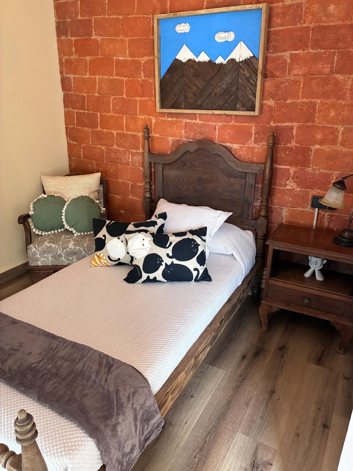 A bed with a wooden headboard, a nightstand with a lamp, and a painting of mountains on the wall.