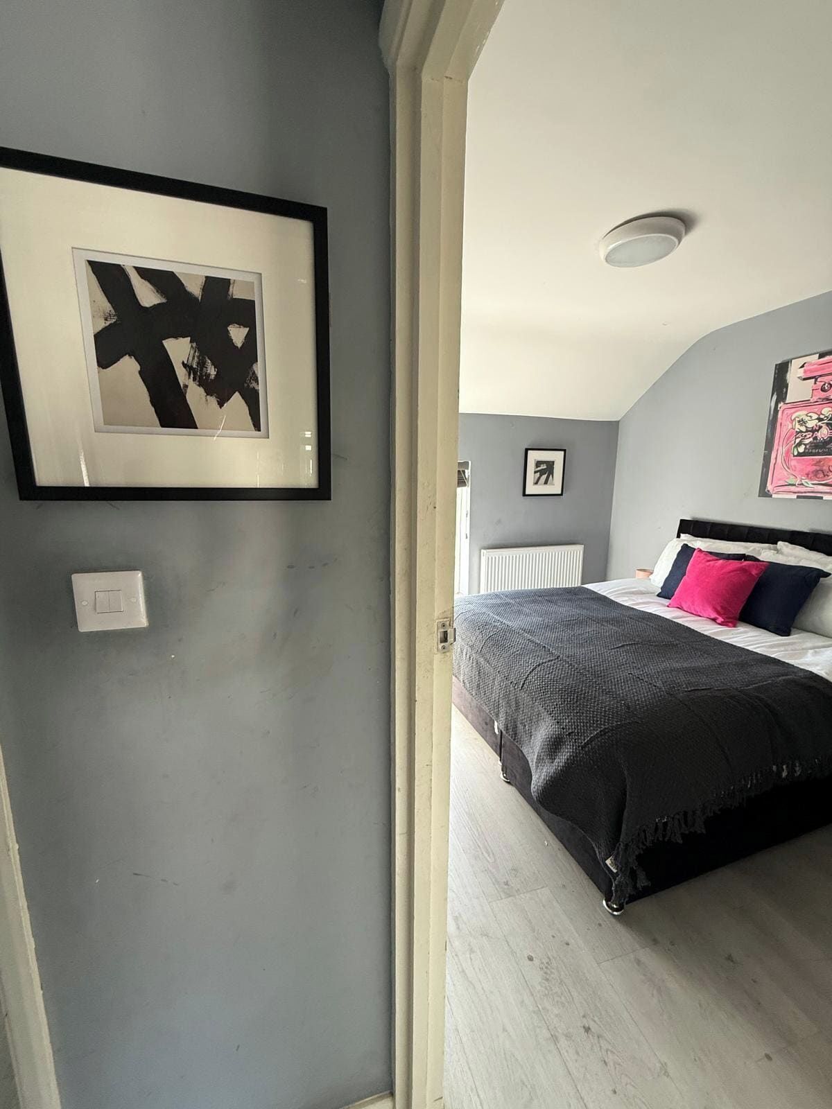 Ein Schlafzimmer mit einem Bett, einem gerahmten abstrakten Gemälde, einem Heizkörper und einer Tür zu einem anderen Raum.