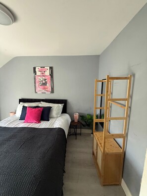 Une chambre à coucher avec un lit, une échelle en bois, une petite table sur laquelle est posée une bougie rose, et un tableau accroché au mur.