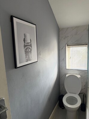 Ein Badezimmer mit Toilette, einem gerahmten Bild an der Wand und einem Fenster mit Jalousien.