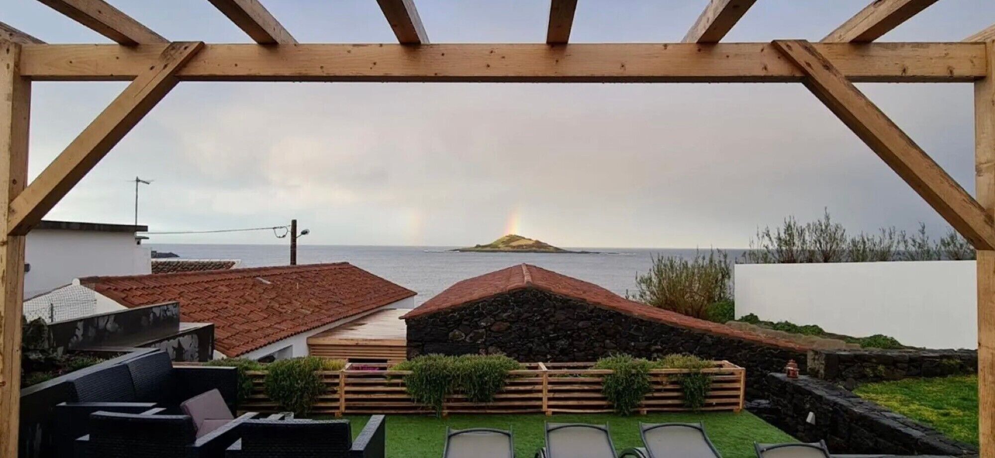 A wooden pergola overlooking a coastal view with a stone structure and a grassy area with chairs.