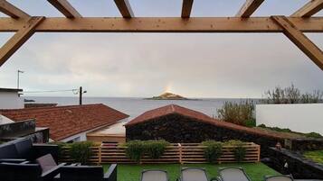 A wooden pergola overlooking a coastal view with a stone structure and a grassy area with chairs.