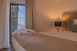 Ein Hotelzimmer mit Bett, Nachttisch mit Lampe und Meerblick durch ein Vorhangfenster.