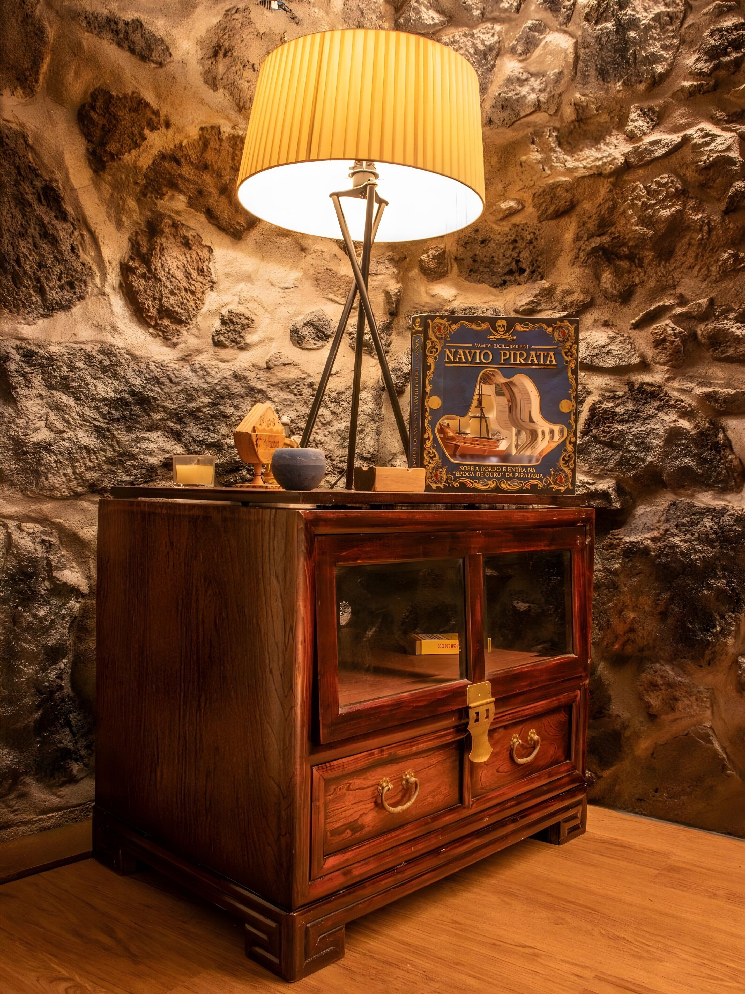 Een houten kast met een glazen vitrine, een lamp en een boek getiteld 'Navio Pirata' tegen een ruwe stenen muur.