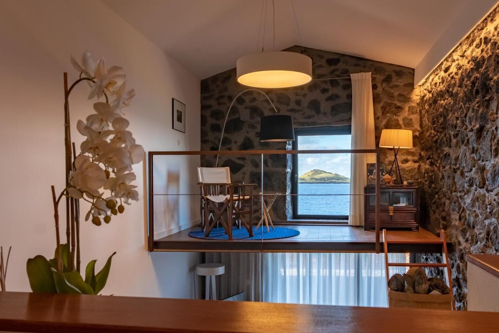 Quarto com vista para o mar, uma escrivaninha de madeira, uma cadeira, um abajur e uma planta.