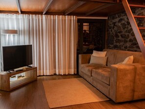 Ein Wohnzimmer mit Fernseher auf einem Holzboden, einem beigefarbenen Sofa, einem großen Fenster mit durchsichtigen Vorhängen und einer Steinwand.
