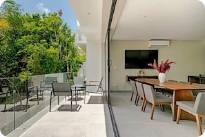 A modern dining area with a large table and chairs, a flat-screen TV mounted on the wall, and a glass railing overlooking a lush garden. - Modern Oasis Near the Beach (Q.R.)