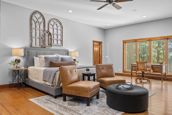 A bedroom with a bed, two chairs, a table, a ceiling fan, and a large window with blinds.