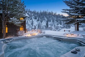 A log cabin with a hot tub surrounded by snow-covered trees and a mountain in the background. - Whispering River Ranch | Hot Tub + Riverfront (Breckenridge)