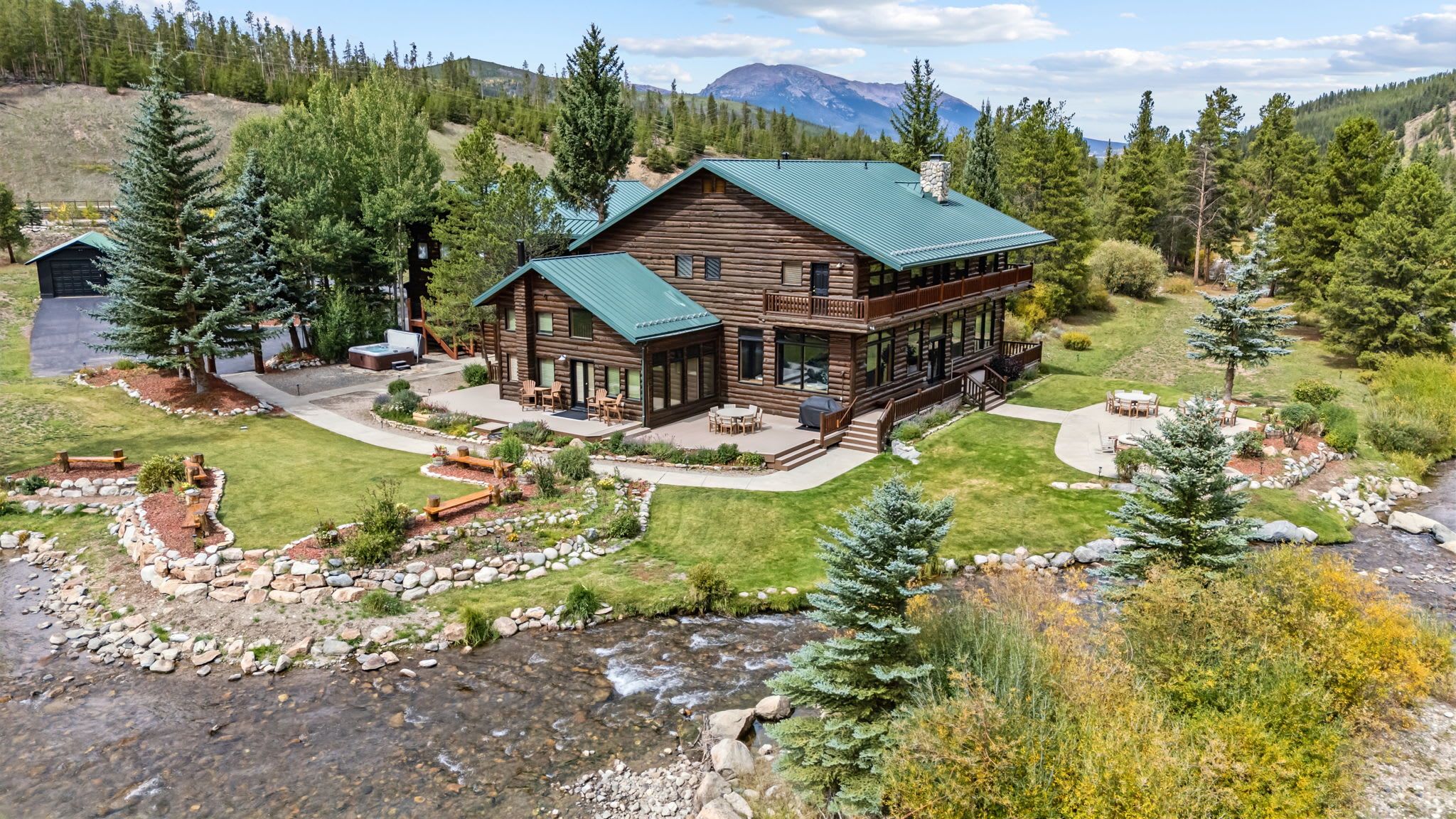 A log cabin with a green roof, surrounded by a well-maintained yard with a patio, a hot tub, and a view of a mountain range.