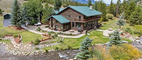 A log cabin with a green roof, surrounded by a well-maintained yard with a patio, a hot tub, and a view of a mountain range.