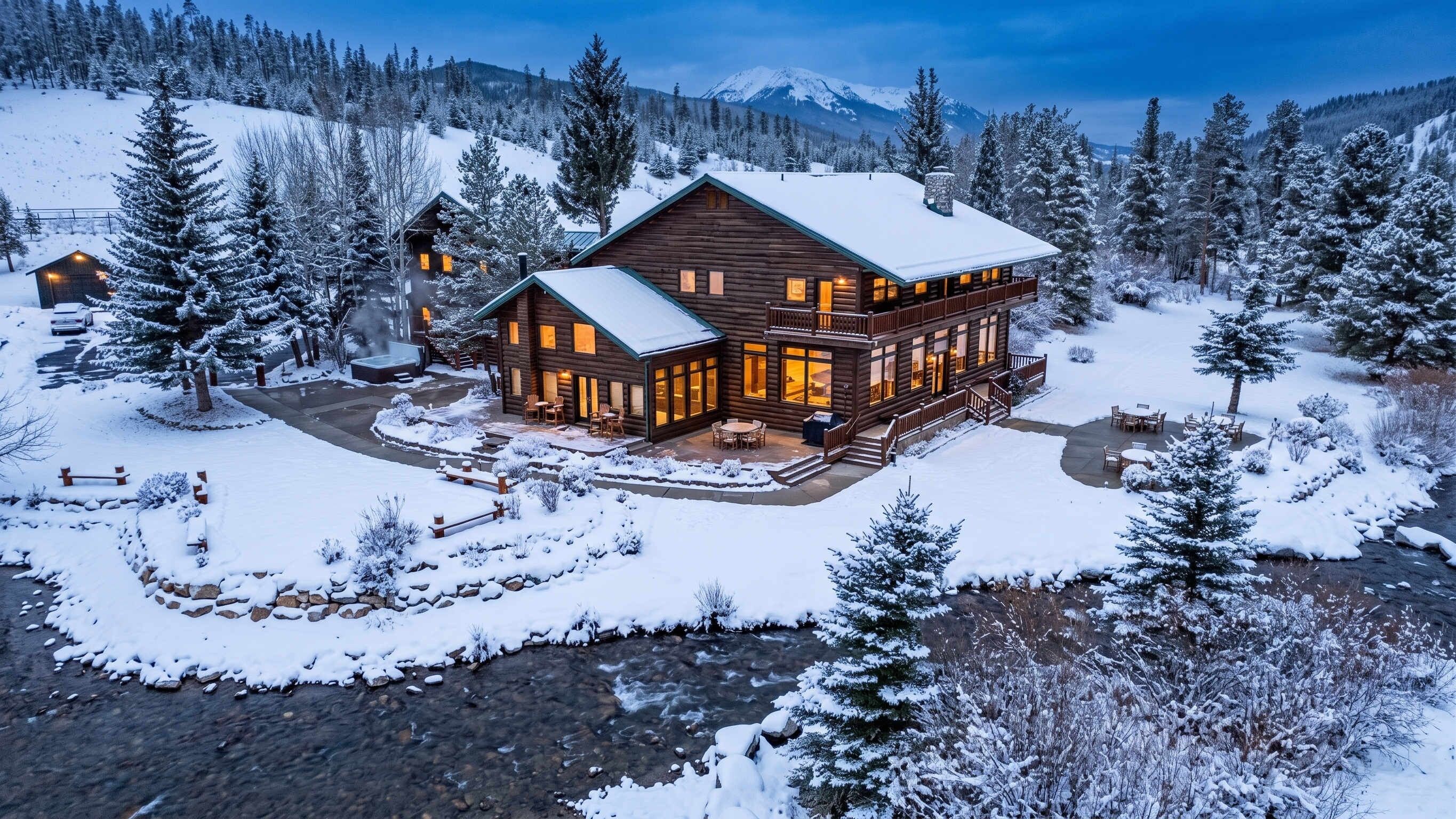 A log cabin with a large deck and a hot tub, surrounded by snow-covered trees and a mountain in the background.