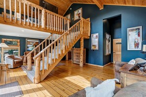 A spacious living area with a wooden staircase, a leather sofa, a coffee table, and artwork on the walls. - Whispering River Ranch | Hot Tub + Riverfront (Breckenridge)
