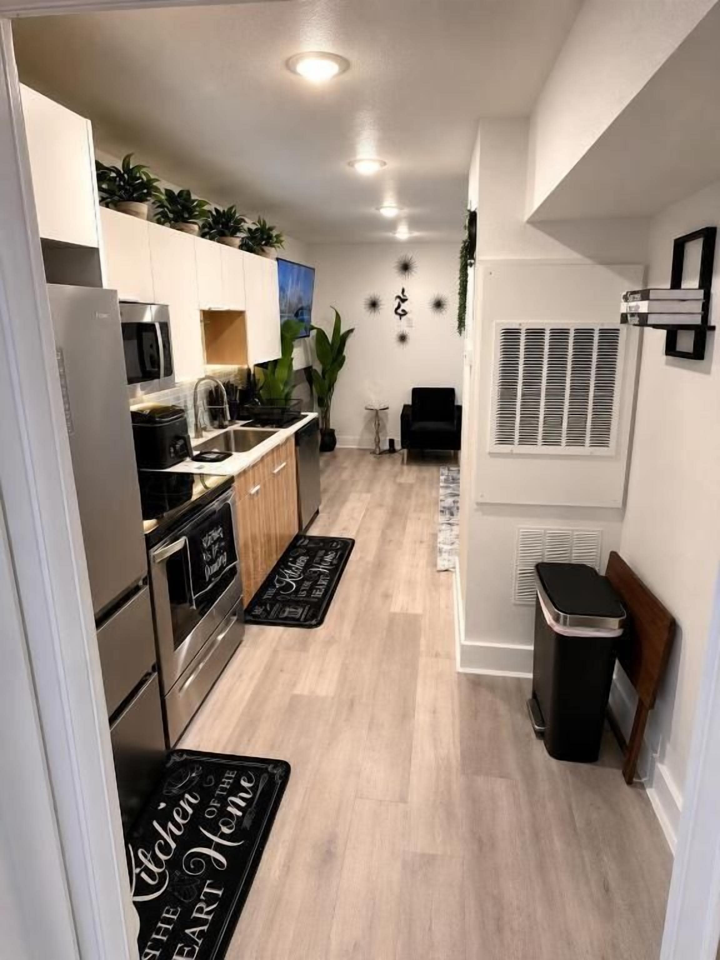 Una cocina estrecha con microondas, estufa y armarios. Hay felpudos negros en el piso y un cubo de basura.