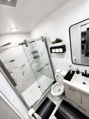 A modern bathroom with a glass shower enclosure, a toilet, a sink with a mirror, and a small shelf with toiletries. - Modern Oasis Near Deep Ellum (Dallas)