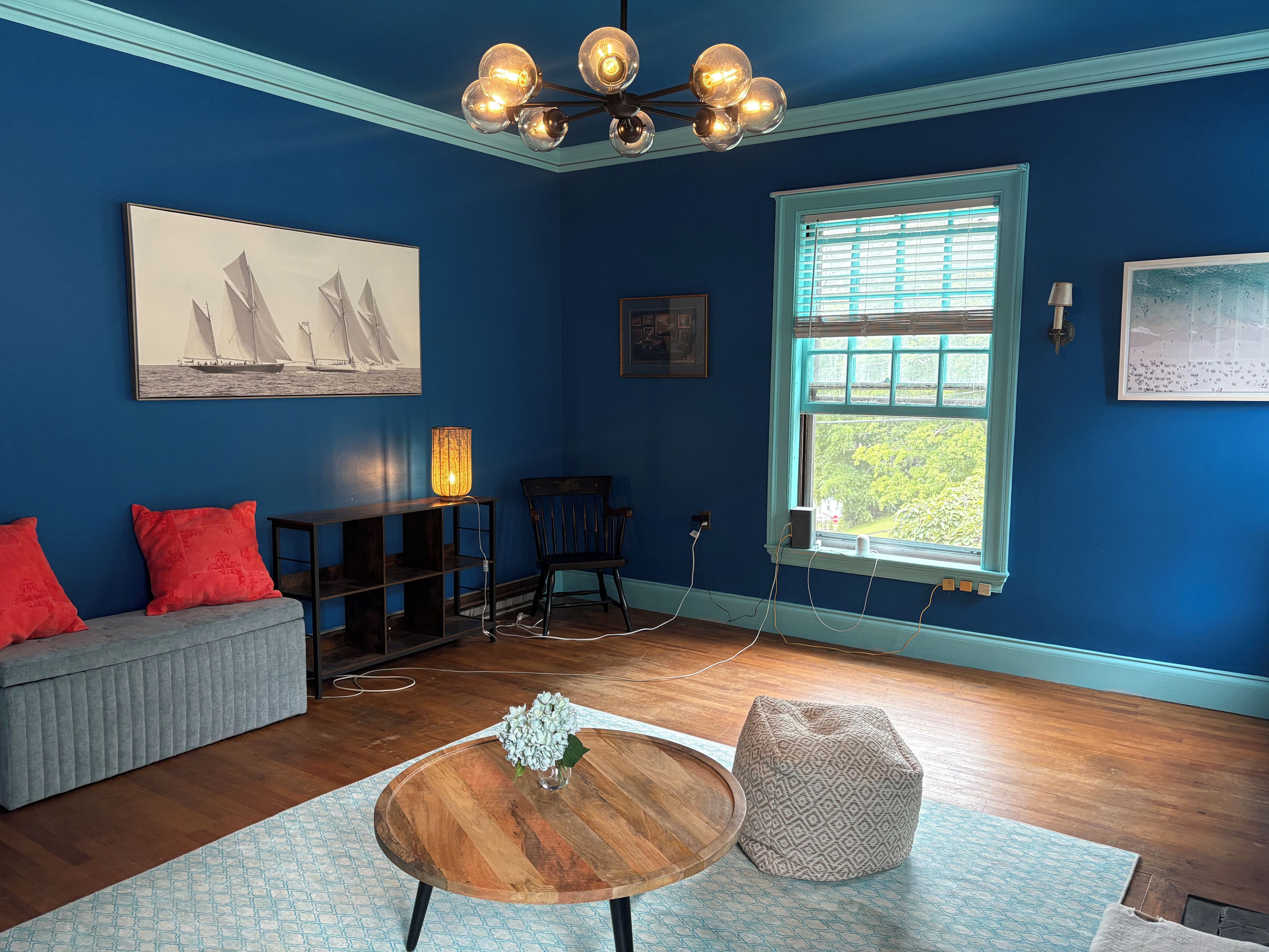 Una stanza con una parete blu, un tavolino da caffè in legno, un pouf decorato, un divano grigio con cuscini rossi e una finestra con persiane.