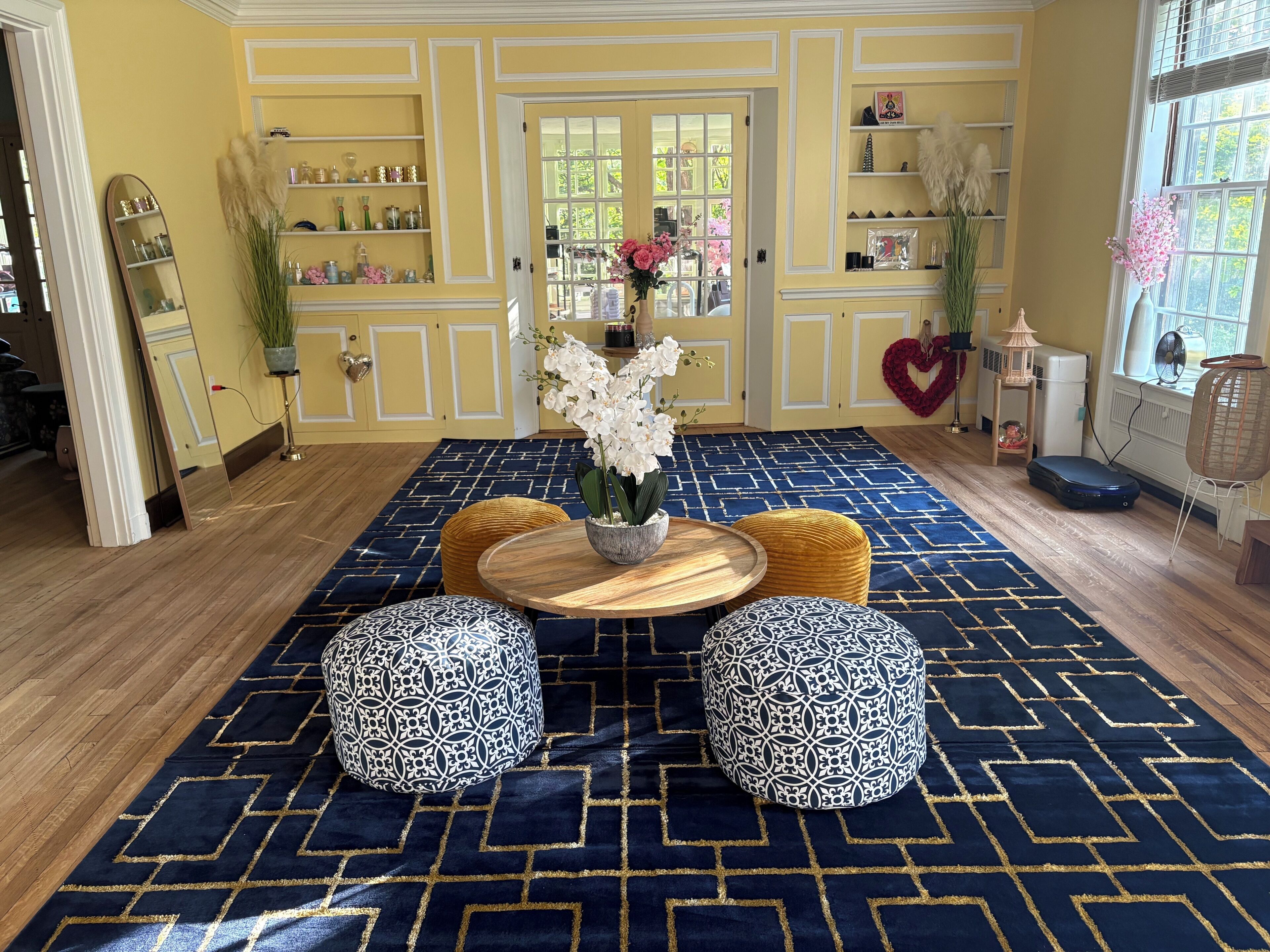 Ein Zimmer mit einem gemusterten Teppich, einem runden Holztisch mit Blumenvase und zwei gemusterten Poufs.