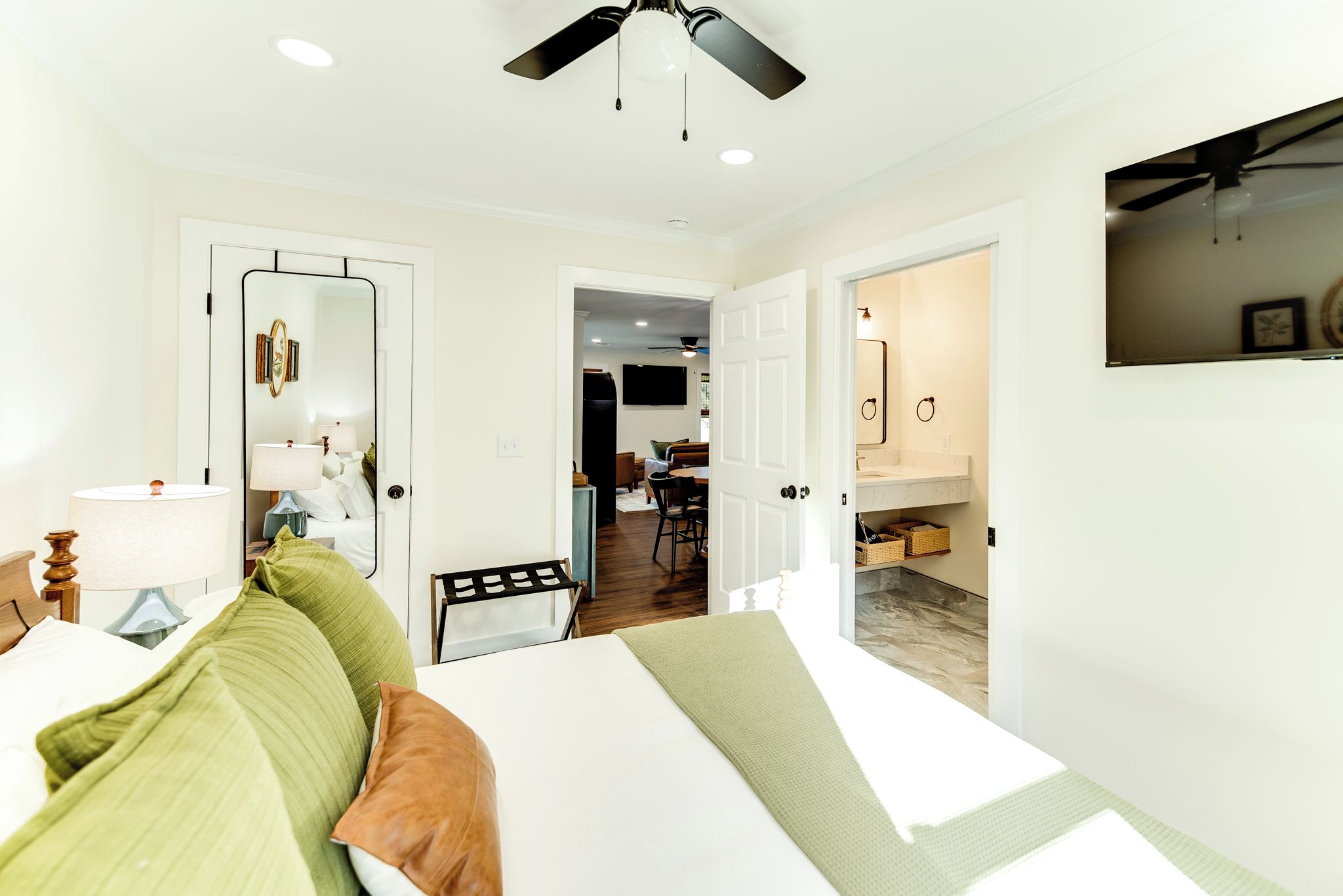 A hotel room with a bed, a desk, a chair, a mirror, a ceiling fan, and a bathroom visible through an open door.