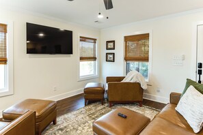 A living room with a flat-screen TV, two windows with blinds, a brown leather sofa, a matching ottoman, and a patterned rug.