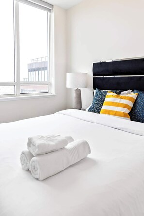 Ein Hotelzimmer mit einem großen Bett, weißer Bettwäsche, einem schwarzen Kopfteil und einem Fenster mit Blick auf Gebäude.