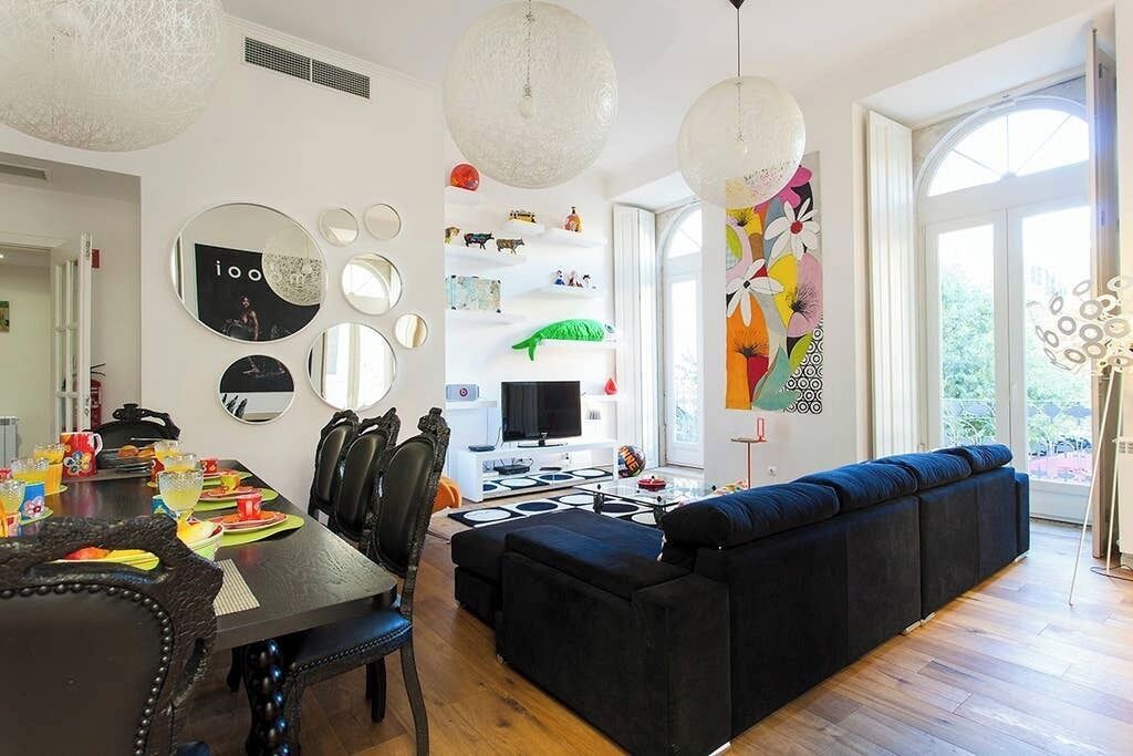 A modern living room with a black sofa, a dining table with chairs, a TV, and a large window.