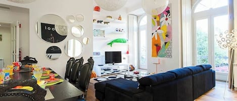 A modern living room with a black sofa, a dining table with chairs, a TV, and a large window.