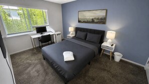 A bedroom with a bed, two bedside tables with lamps, a desk with a computer, and a window with blinds.