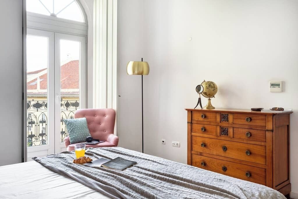 Ein Schlafzimmer mit Bett, Schminktisch, Stuhl und einem Fenster mit Blick auf Gebäude.