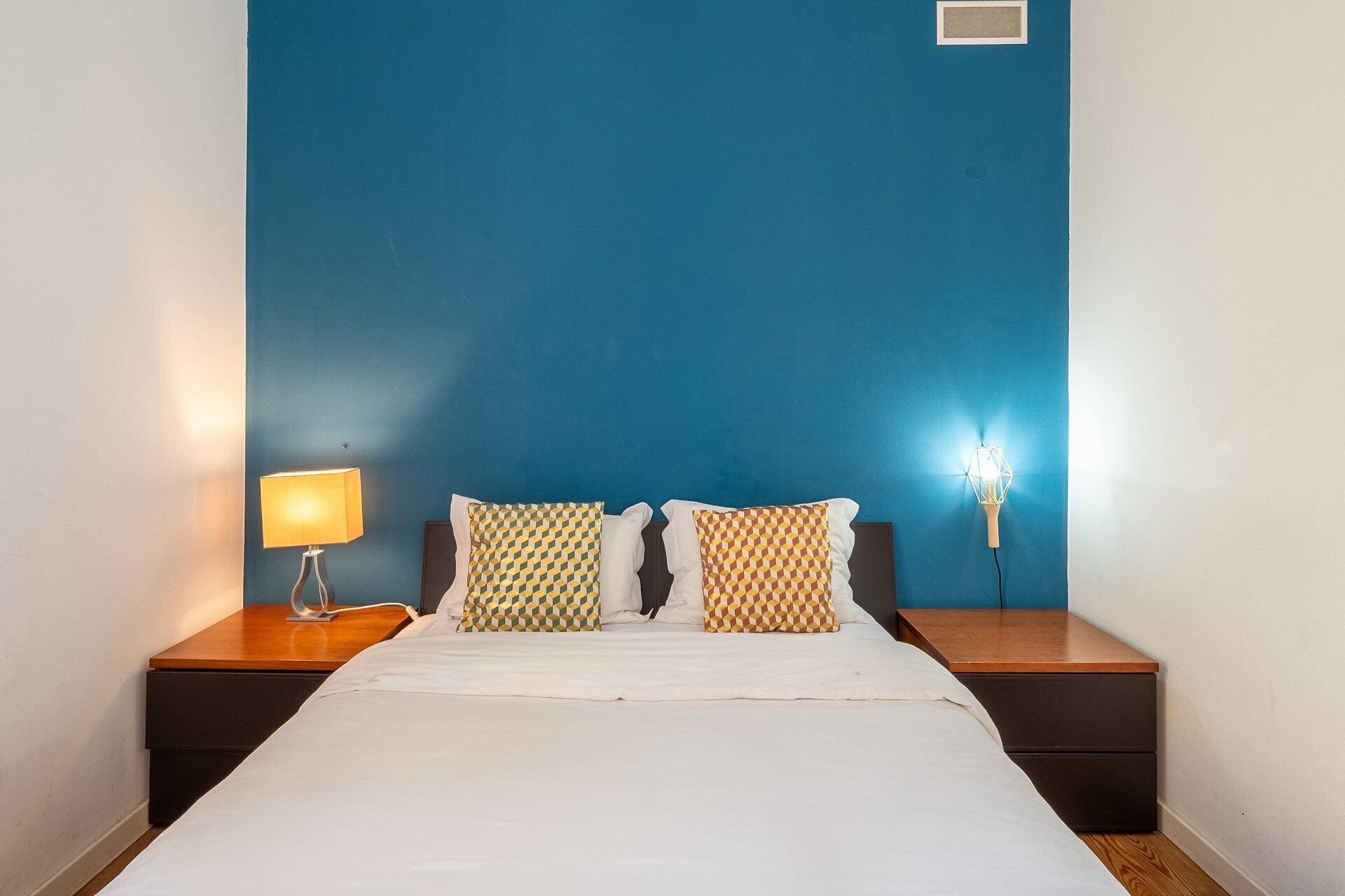A hotel room with a bed, two patterned pillows, a nightstand with a lamp, and a wall-mounted light fixture.