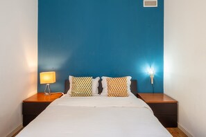 Habitación de hotel con cama, dos almohadones estampados, mesita de noche con lámpara y aplique de pared.