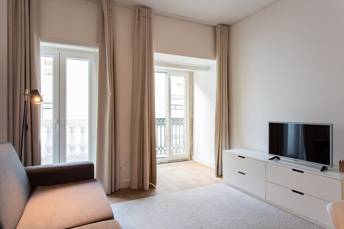 Un salon moderne avec un canapé, un téléviseur à écran plat posé sur un meuble blanc et un balcon agrémenté de rideaux.