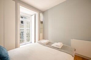 A bedroom with a bed, a radiator, a window with a view of a balcony, and a door leading to another room. - GuestReady - Fantastic flat near S. Jorge Castle (Lisbon)