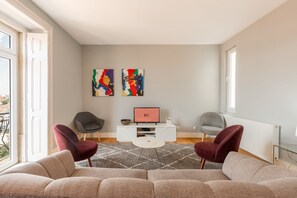 A modern living room with a sofa, two armchairs, a coffee table, and a TV on a white cabinet. There are abstract paintings on the wall and a window with a view. - GuestReady - Fantastic flat near S. Jorge Castle (Lisbon)