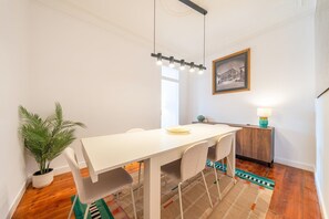 Un coin repas avec une table blanche, des chaises, un luminaire suspendu, un tableau encadré au mur et une plante en pot.