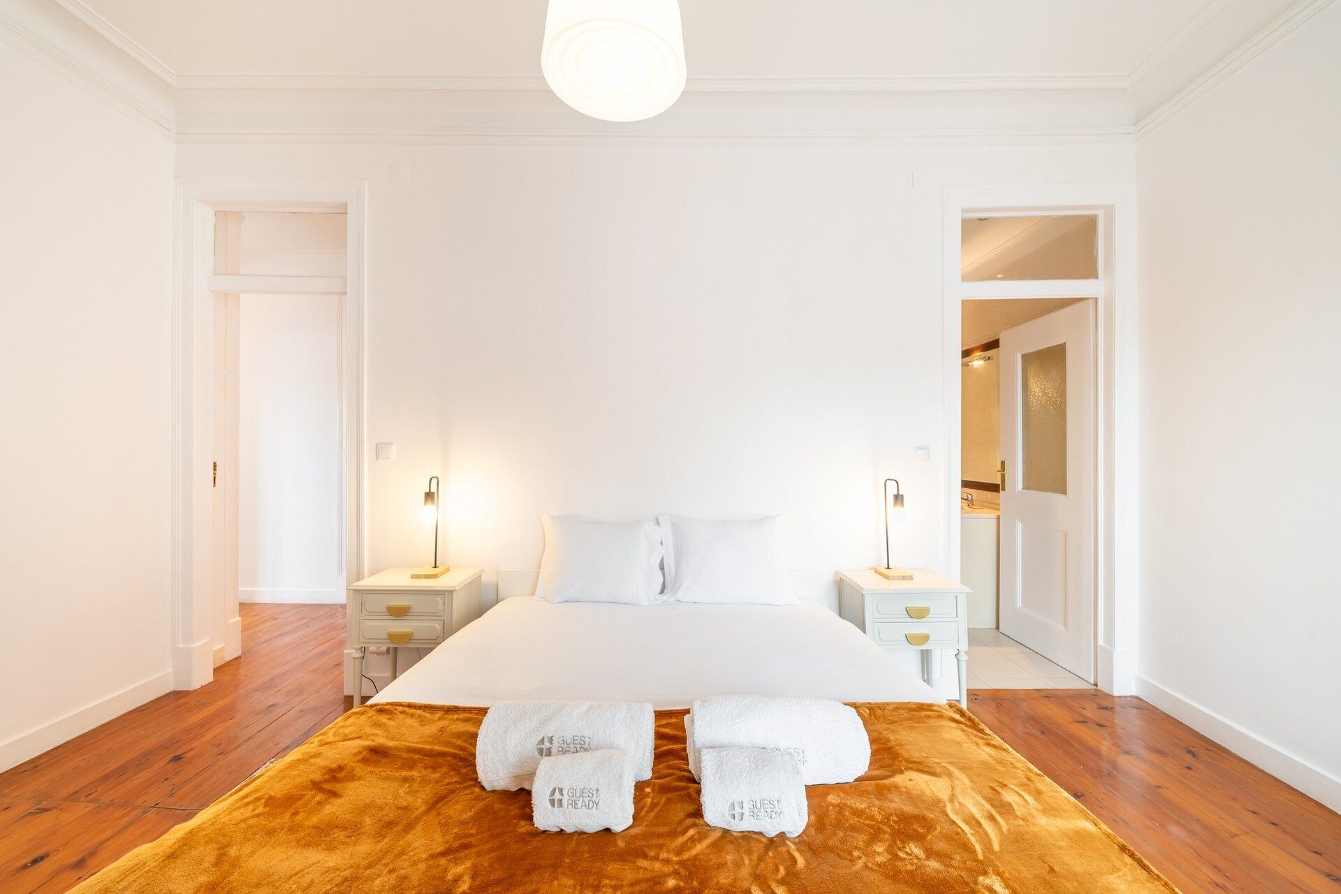 A hotel room with a large bed, two bedside tables, a nightstand, a wooden floor, and a door leading to a bathroom.