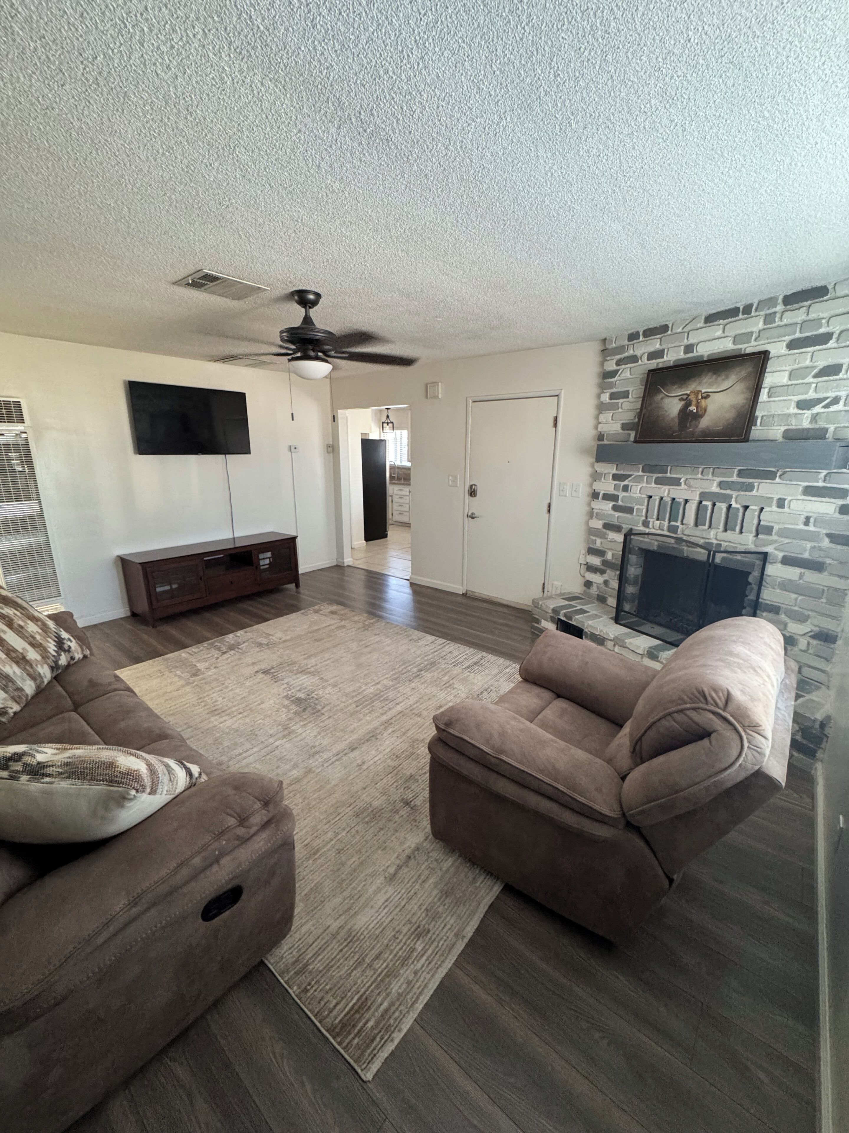 A living room with a flat-screen TV, a ceiling fan, a stone fireplace, and a door leading to another room.