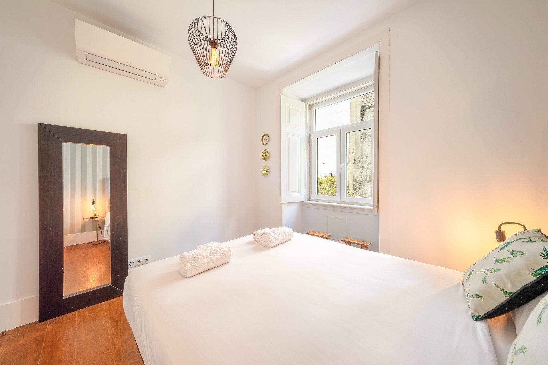 A bedroom with a large bed, a mirror, a window, and a hanging light fixture.