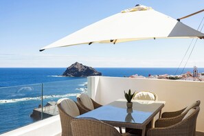 Ein Balkon mit Korbmöbeln, einem Glastisch und einem weißen Sonnenschirm mit Blick auf das Meer und eine felsige Insel.