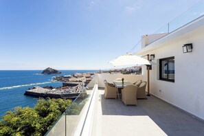 Ein Balkon mit Tisch und Stühlen, der auf eine Küstenlandschaft mit Felsvorsprung und blauem Himmel blickt.